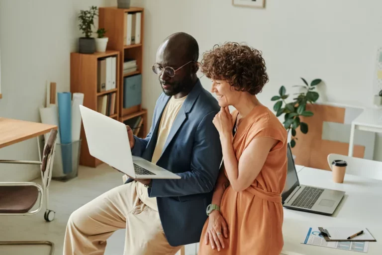Man and woman sitting in an open plan office, reading laptop screen together